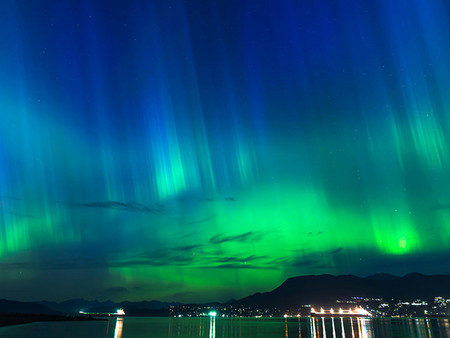 Kosmische Lichter erhellen den Nachthimmel in Grün und Blau, reflektiert von einem stillen See im Vordergrund; dahinter liegen eine Stadt und Berge bei Nacht