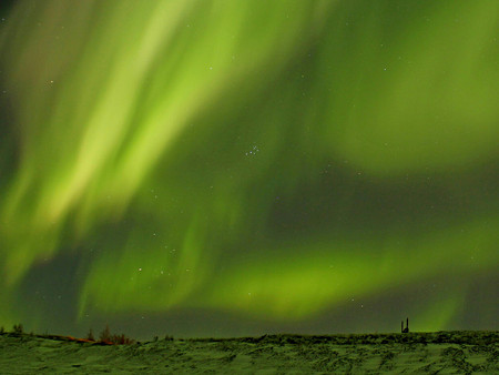 Kosmische Lichter erhellen den Nachthimmel in Grün und Gelb, darunter eine ruhige, verschneite Landschaft