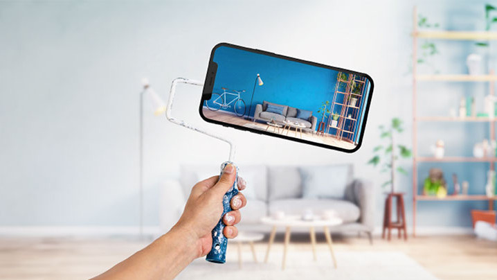 Person hält Farbrolle mit Handy als Rolle in Wohnzimmer – auf dem Handy das umgestaltete Wohnzimmer sichtbar. Spectrum Anwendung.