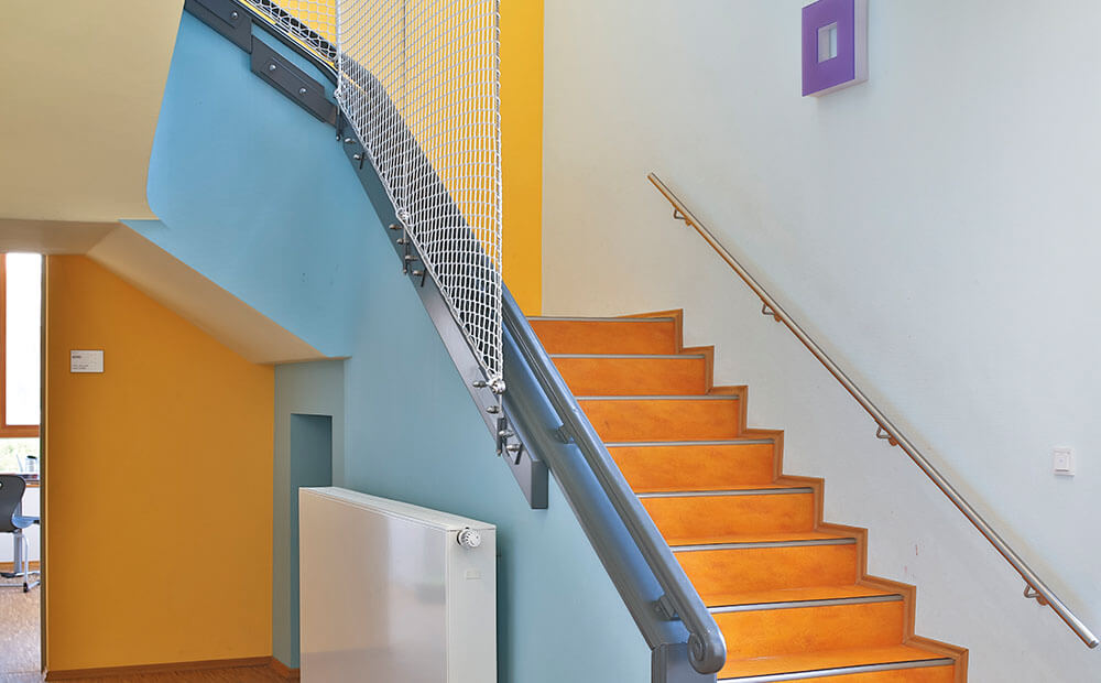 Blau trifft Orange: Im Treppenhaus des Rosbacher Altbaus, einer ehemaligen Schule, regte die vorgegebene Wandbeleuchtung dazu an, die geschwungene Brüstung in Blautönen anzulegen. Der Gesamteindruck: frisch, belebend und harmonisch. Hier fühlt sich jedes Kind gut aufgehoben. 