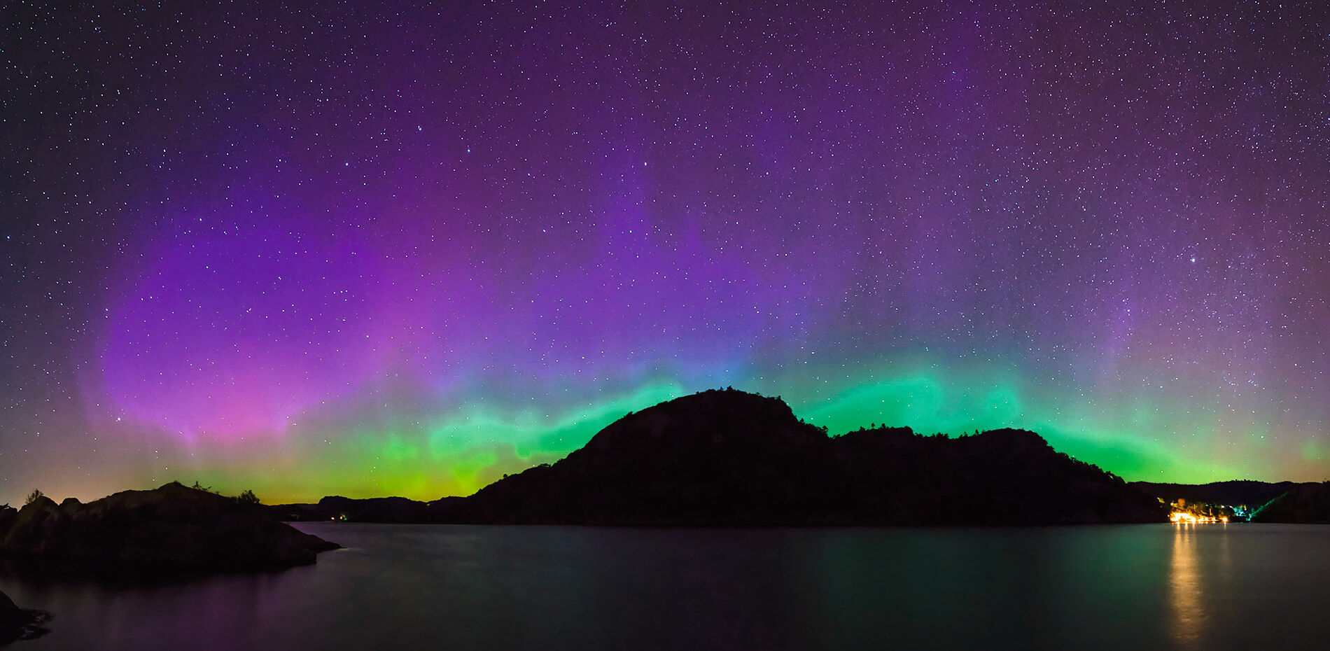 Kosmisch leuchtende Polarlichter in Grün und Lila am Nachthimmel, mit einem stillen See im Vordergrund, der die Farben reflektiert, und Bergen im Hintergrund