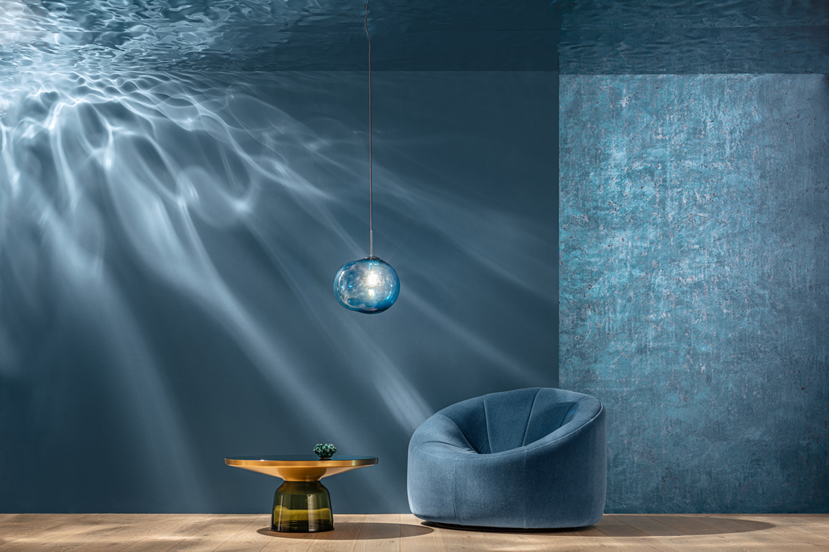 Ein gemütlicher, blauer Sessel steht vor einer blauen Wand mit sanften Lichtstrahlen. Neben dem Sessel befindet sich ein runder Tisch mit einer goldenen Oberfläche und einem grünen Dekorationsobjekt darauf. Über dem Sessel hängt eine gläserne Pendelleuchte in Blau.